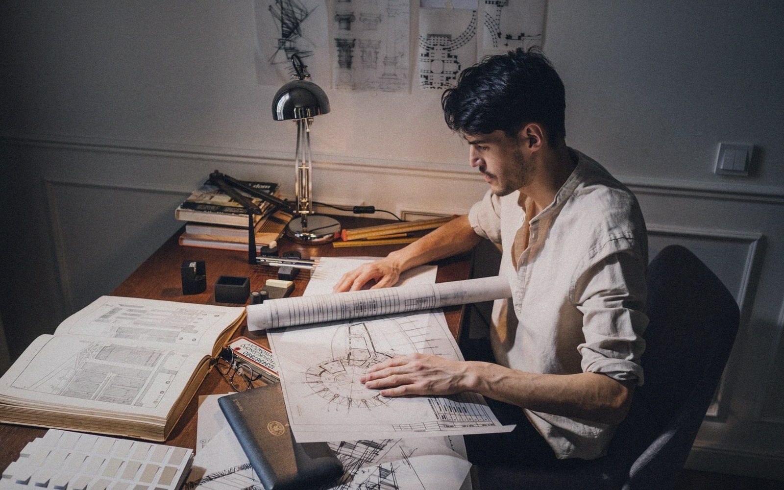 A draughtsman at a desk reading a large drawing, reference books open beside him